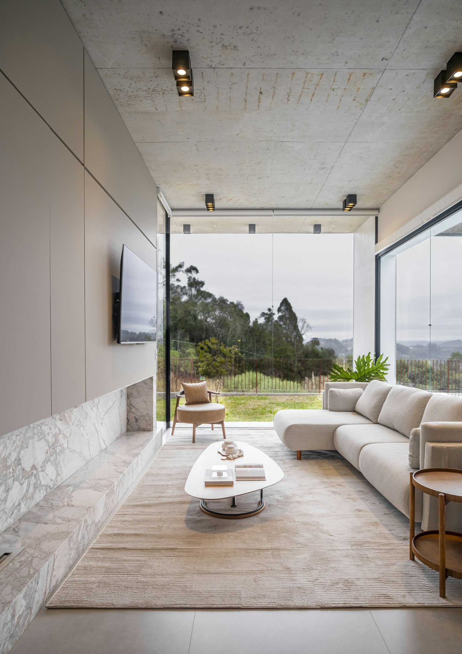 Invisible grill design for balcony: living room opening onto a balcony through a full-height glass front, thin vertical railing line barely visible against the trees